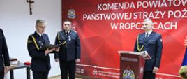 Mł. kpt. Marcin Pasko odczytuje decyzje o zwolnieniu ze służby st.og. Łukasza Nowiecia, strazacy w umundurowaniu wyjsciowym na tle logo KP PSP