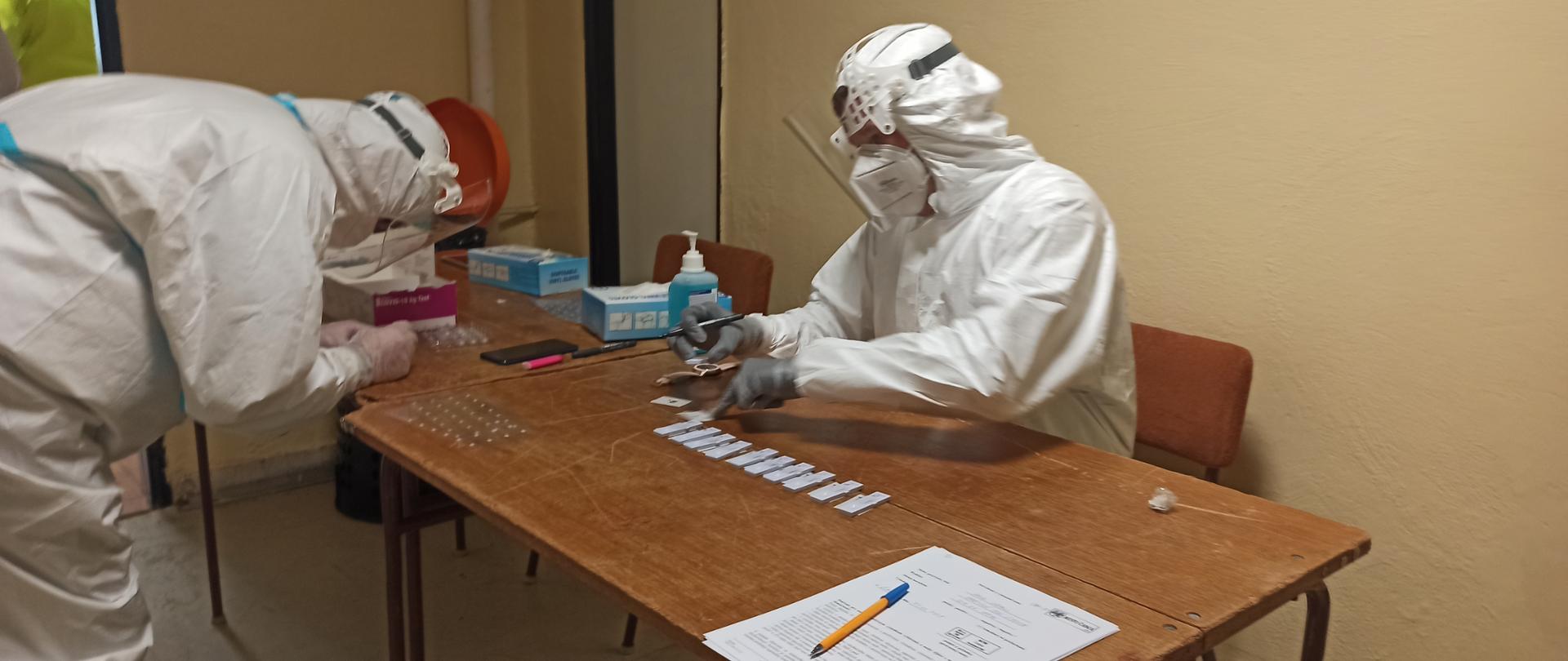 Strażacy w kombinezonach ochronnych na stanowisku pracy w trakcie opisywania pobranych próbek