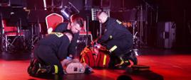 Na zdjęciu widocznych trzech strażaków z Komendy Powiatowej Państwowej Straży Pożarnej w Gorlicach w ubraniach koszarowych czarnych w trakcie pokazu kwalifikowanej pierwszej pomocy na scenie 32 Finału Wielkiej Orkiestry Świątecznej Pomocy na hali Ośrodka Sportu i Rekreacji w Gorlicach.