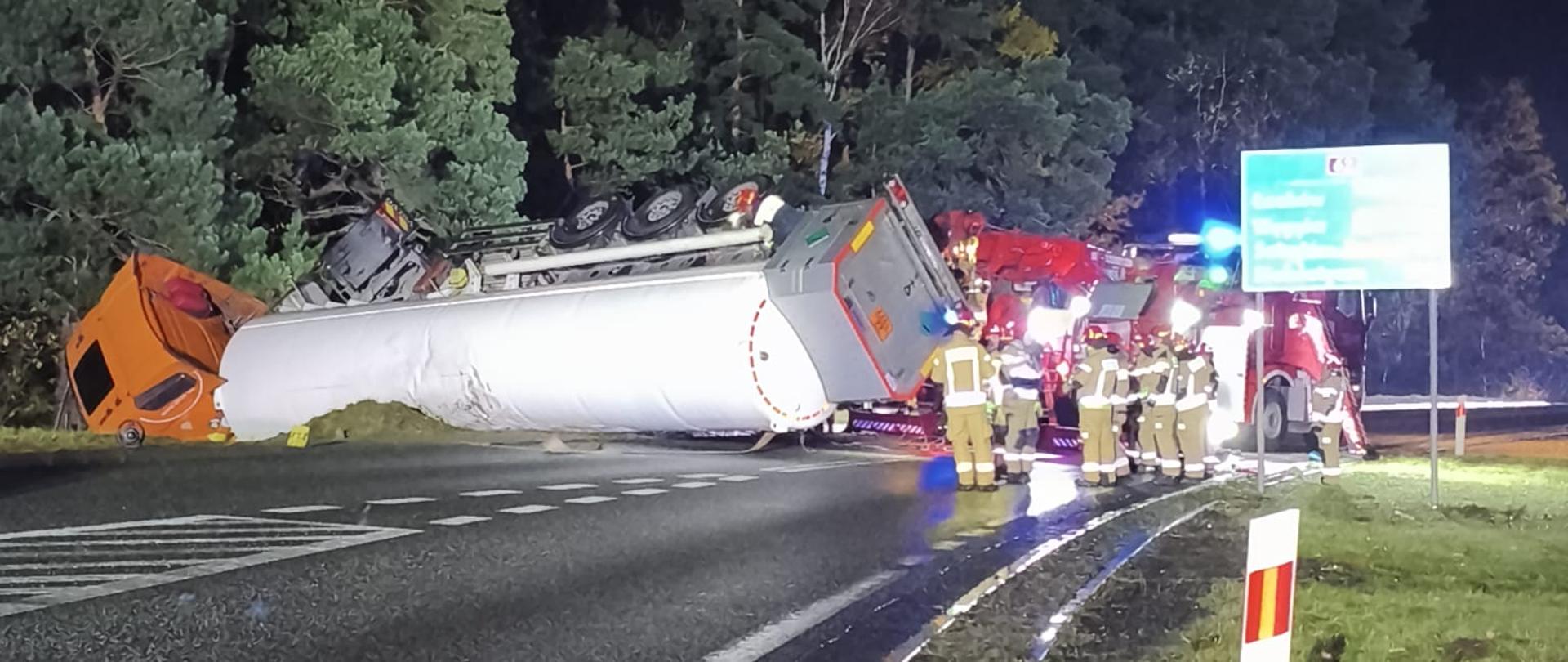 Przewrócona biała cysterna na drodze w czasie nocy. Widoczni strażacy w ubraniach specjalnych i ciężki samochód ratownictwa technicznego.