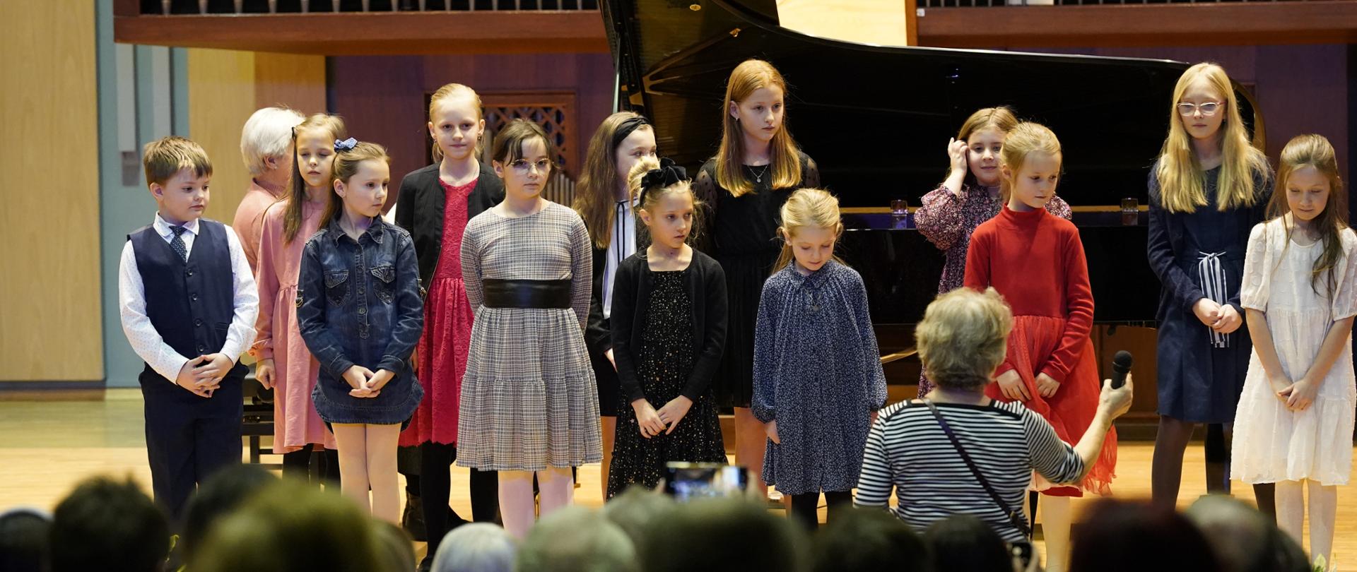 Uczniowie stoją w dwóch rzędach na scenie sali koncertowej. Na pierwszym planie publiczność. Przed uczniami stoi nauczycielka trzymająca w dłoni mikrofon. Za uczniami fortepian. Przy fortepianie gra Pani akompaniatorka, w tle organy.