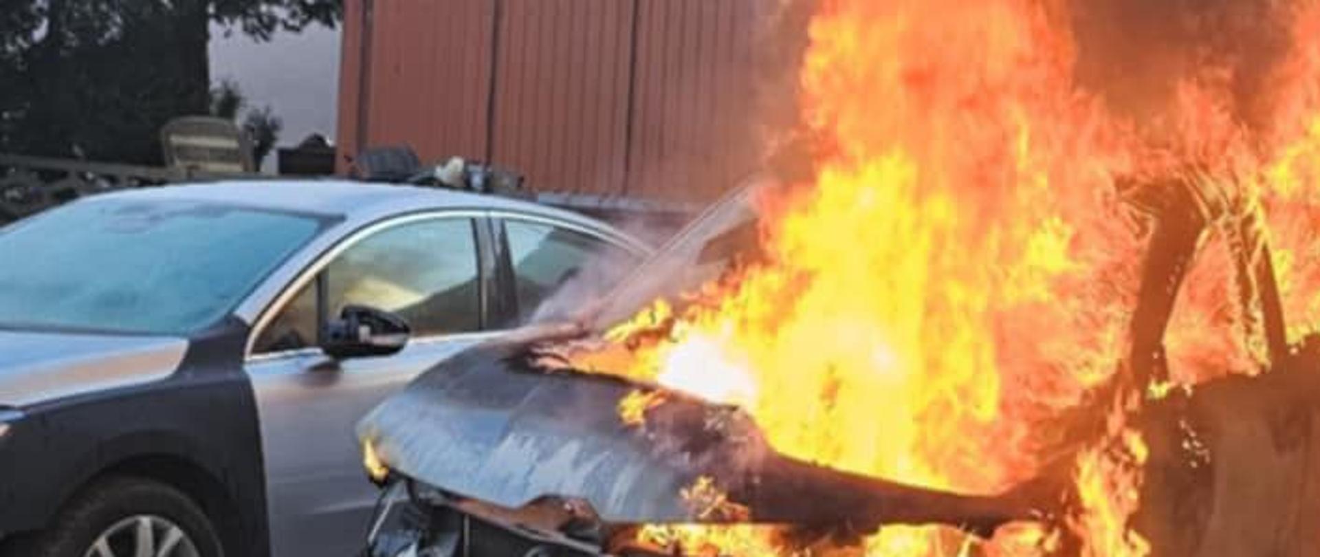 Na zdjęciu palący sie samochód osobowy w całej objętości. Widać wychodzące płomienie z bryły pojazdu. Ogień zagraża sąsiedniemu pojazdowi