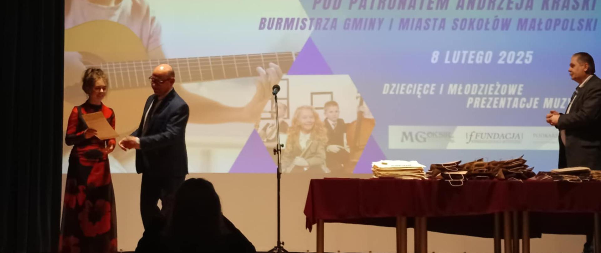 Scena, a na niej dwie osoby. Dziewczyna w długiej czarnej sukni w czerwone maki odbiera dyplom od pana z jury. N scenie stół na którym poukładane są torebki z nagrodami.