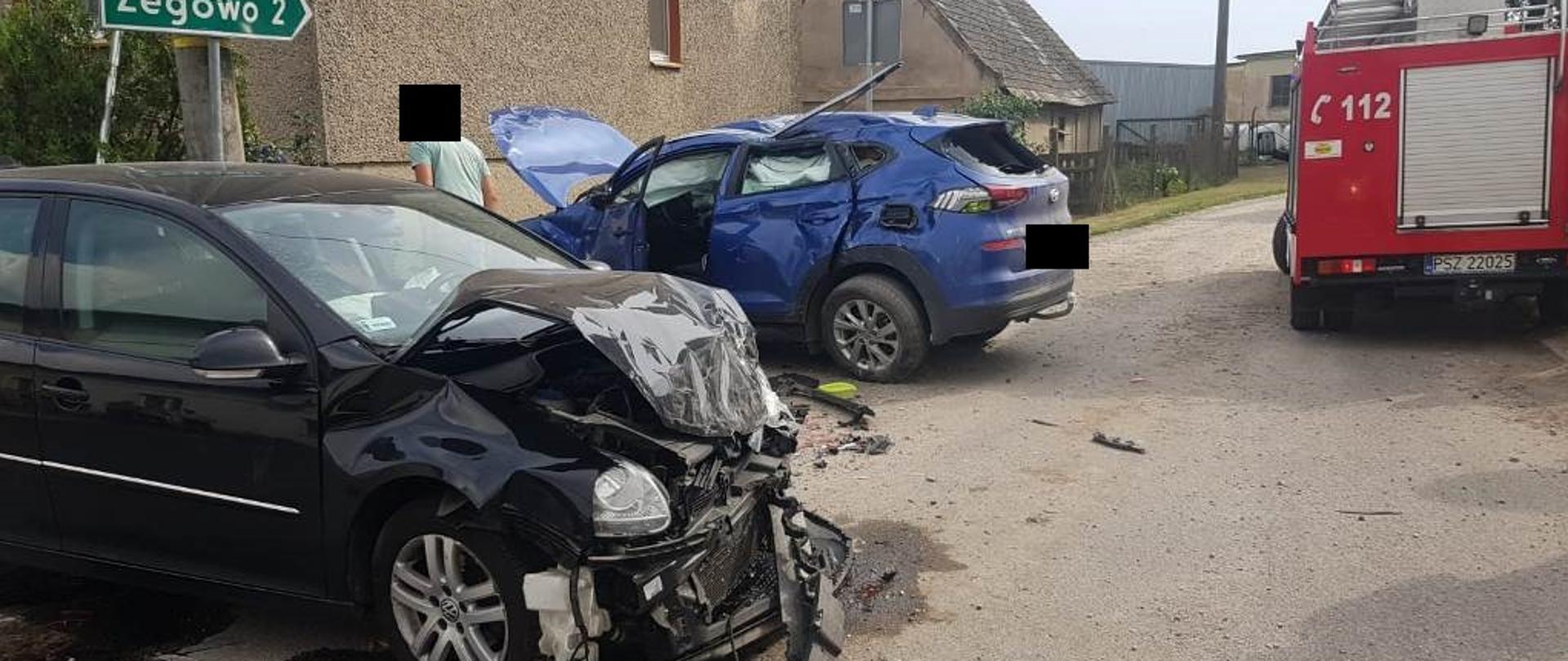 Czarny samochód ma uszkodzony przód, obok znajduje się niebieski samochód z podniesioną maską, ma uszkodzony lewy bok, dach i tył, w pobliżu jest samochód strażacki