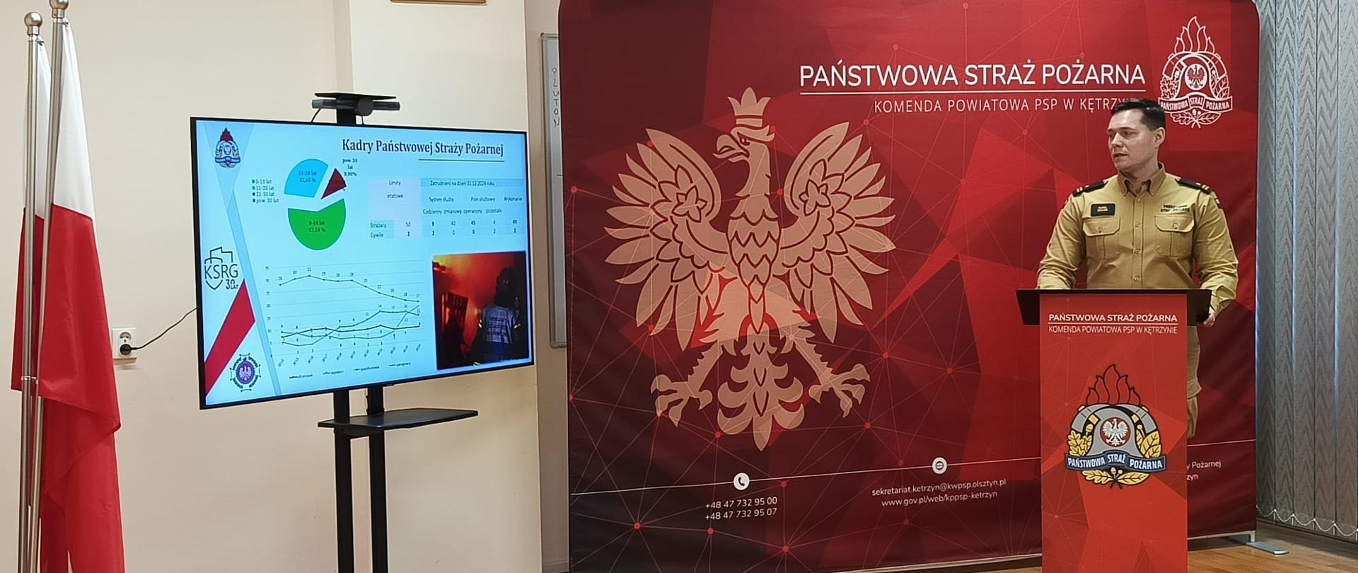 Na zdjęciu widać strażaka w żółtym ubraniu służbowym stojącego przed mównicą. Za nim widać czerwony baner z logo i napisem Państwowa Straż Pożarna.