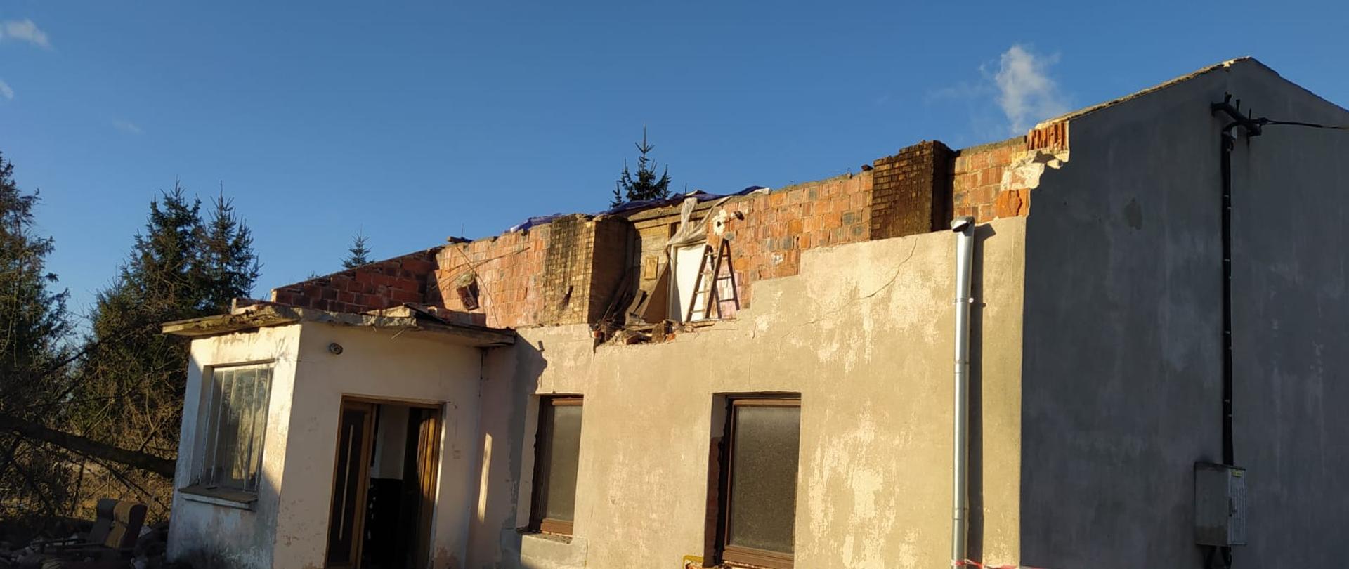 Na zdjęciu widać uszkodzony budynek. Budynek nie ma dachu. Widać odsłonięte kominy z cegły. Ściany budynku szare. Przed budynkiem gruz. Na dalszym planie drzewa iglaste w kolorze zielonym. Niebo bezchmurne, kolor niebieski.