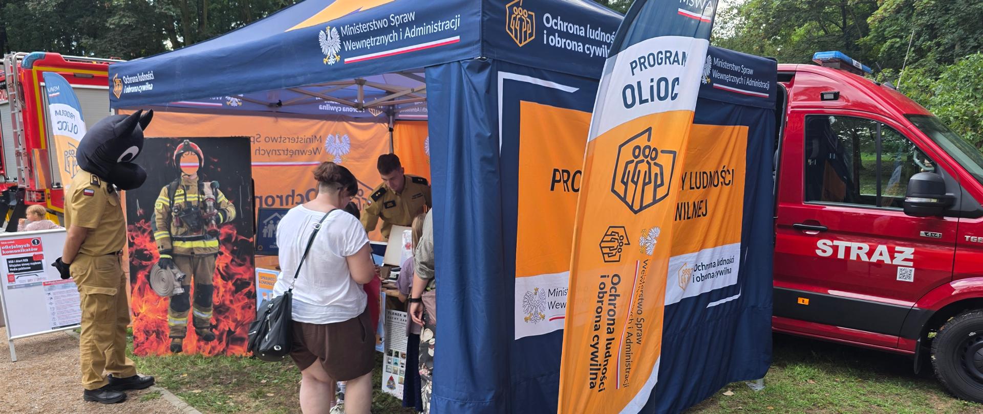 Zdjęcie przedstawia plenerowe stoisko edukacyjno-informacyjne zorganizowane przez Ministerstwo Spraw Wewnętrznych i Administracji (MSWiA) oraz Państwową Straż Pożarną, skupione wokół tematyki ochrony ludności i obrony cywilnej.
Główne elementy na zdjęciu:
Namiot ekspozycyjny: Niebiesko-pomarańczowy namiot z godłem Polski oraz oficjalnymi napisami „Ministerstwo Spraw Wewnętrznych i Administracji” oraz „Ochrona ludności i obrona cywilna”. Obok stoją flagi wideo (beachflagi) z logo programu OLiOC.
Postać w maskotce: Po lewej stronie stoi osoba w mundurze strażackim (lub służb podległych MSWiA) z nałożoną głową czarnej maskotki (psa/kota), rozmawiająca z odwiedzającymi.
Atrakcje dla dzieci i odwiedzających:
Fotostand (ścianka do zdjęć) z wizerunkiem strażaka na tle ognia z wyciętym otworem na twarz.
Stoisko z materiałami edukacyjnymi, przy którym strażacy/funkcjonariusze w piaszczystych mundurach służbowych rozmawiają z odwiedzającymi (kobieta w białej bluzce i dzieci).
Pojazdy pożarnicze: Po prawej stronie widoczny jest czerwony furgon strażacki (Volkswagen Crafter/MAN TGE) z napisem STRAŻ, a w tle po lewej stronie fragment większego wozu gaśniczego.
Otoczenie: Wydarzenie odbywa się na świeżym powietrzu, na terenie zielonym (park lub plac festynowy) w otoczeniu drzew.