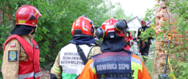 Grupa ratowników w hełmach i kombinezonach stoi przy częściowo zawalonej ceglanej konstrukcji otoczonej zielenią. Na kamizelkach widoczne są napisy „Kierujący Działaniem Ratowniczym” i „Dowódca SGPR”. Wokół znajdują się liny i sprzęt wykorzystywany podczas działań.
