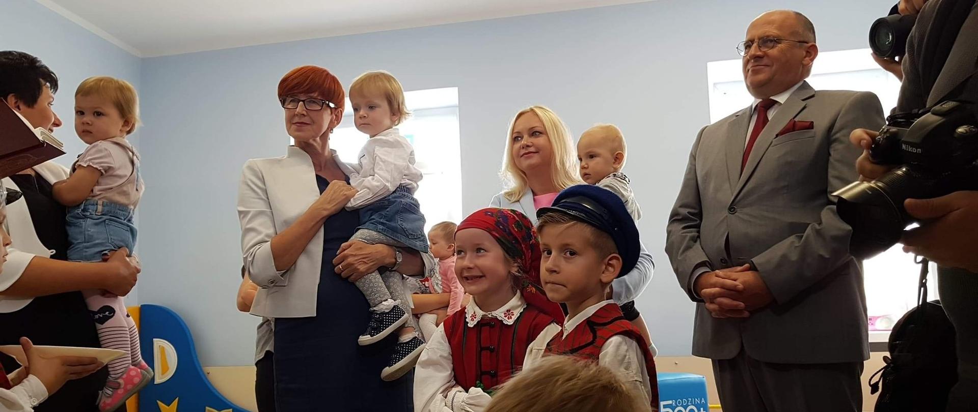 Otwarcie pierwszego żłobka w Łęczycy. Minister Elżbieta Rafalska trzyma na rękach dziewczynkę. Towarzyszy jej wojewoda łódzki, dyrektor żłobka. W tle dzieci, które uczestniczą w wydarzeniu.