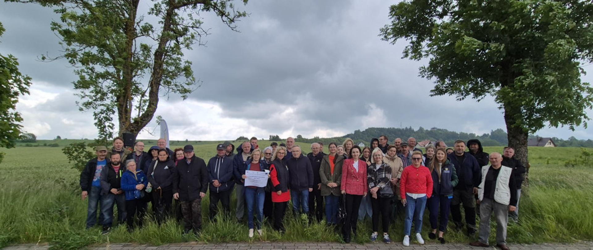 Grupa osób pozuje do zdjęcia. Stoją na polu między dwoma drzewami przed chodnikiem