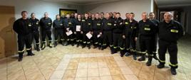 Recertyfikacja kpp. Korytarz w komendzie PSP w Rawiczu. Na tle ściany stoją strażacy egzaminowani w pierwszej grupie, z zaświadczeniami w rękach i w towarzystwie komisji egzaminacyjnej.