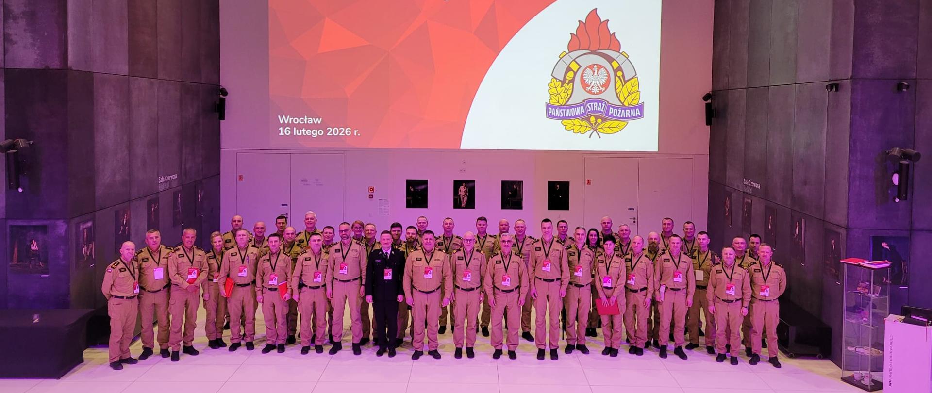 Wspólne zdjęcie uczestników narady rocznej kadry kierowniczej PSP woj. dolnośląskiego – kilkudziesięciu funkcjonariuszy w umundurowaniu pozuje pod ekranem z nazwą wydarzenia.