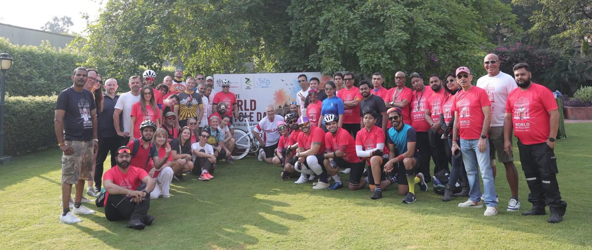 Grupa uczestników rajdu rowerowego pozuje do wspólnego zdjęcia w ogrodzie Rezydencji Ambasadora RP po zakończeniu przejazdu z okazji Światowego Dnia Roweru. Większość osób ma na sobie czerwone, białe lub czarne koszulki wydarzenia, a w tle widoczny jest duży baner z grafiką rowerzysty i zabytków Nowego Delhi.