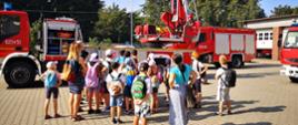 Zdjęcie przedstawia grupę dzieci, które przyglądają się jak strażacy prezentują pracę drabiny mechanicznej.