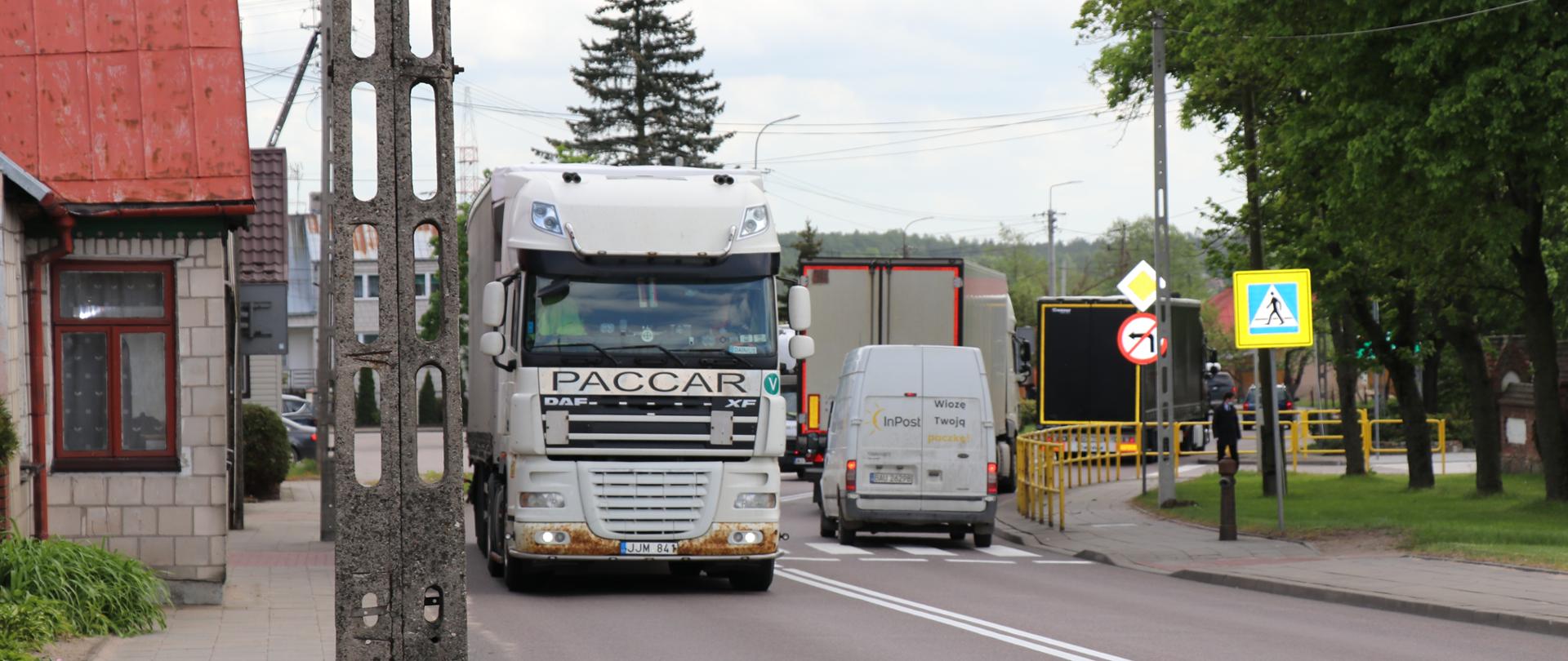 Asfaltowa ulica , na pierwszym planie ciężarówka przodem, na drugim pasie samochód dostawczy tyłem, przed nim kolejne ciężarówki 