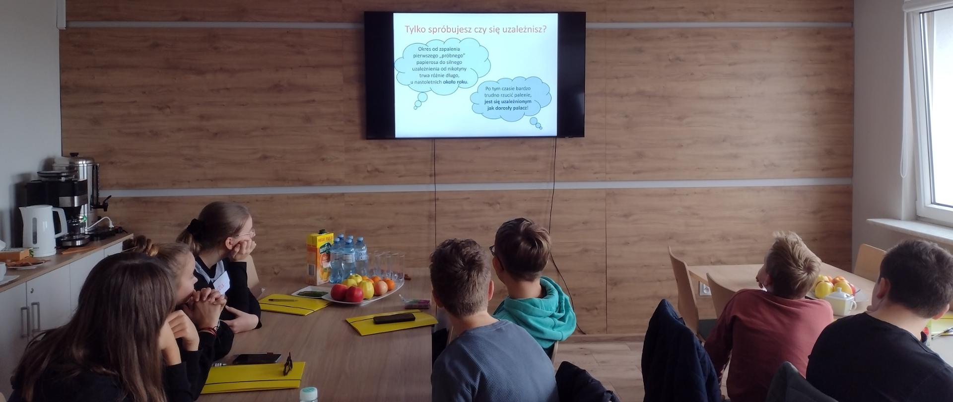 Uczniowie siedzą przy stołach i oglądają prezentację multimedialną wyświetlaną na dużym ekranie