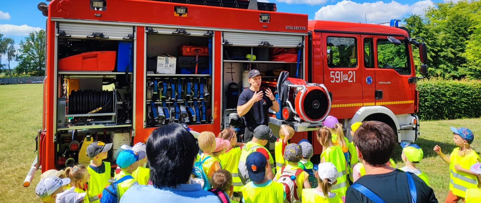 Widać dzieci przed samochodem strażackim, strażak omawia dzieciom działania