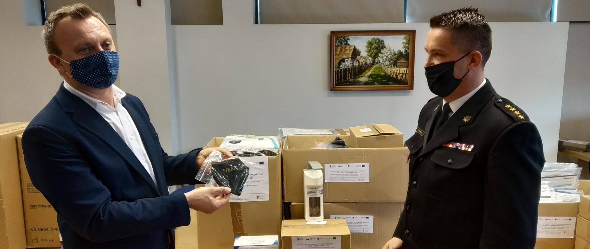 Zdjęcie przedstawia Starostę Marcina Piwnika, który prezentuje maseczkę z napisem straż, Po prawej komendant powiatowy PSP w mundurze wyjściowym patrzy w kierunku starosty. Za nimi na stole stoją pudełka ze środkami ochrony indywidualnej.