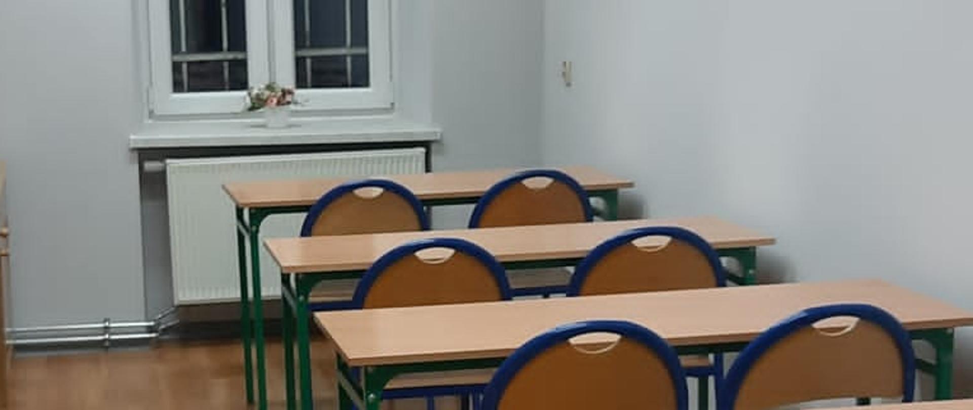 Zdjęcie przedstawia pomieszczenie, w której znajduje się czytelnia. Widoczny jest rząd podwójnych ławek wraz z krzesłami. Na zdjęciu widoczne jest także okno oraz wiszące lampy.