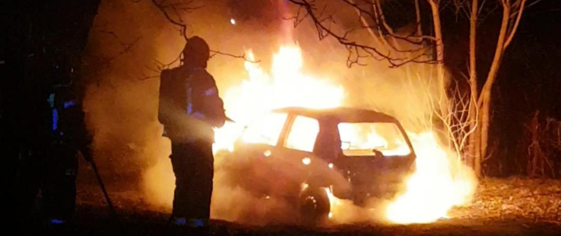 Zdjęcie przedstawia strażaka gaszącego pożar samochodu osobowego. Samochód objęty jest cały ogniem.