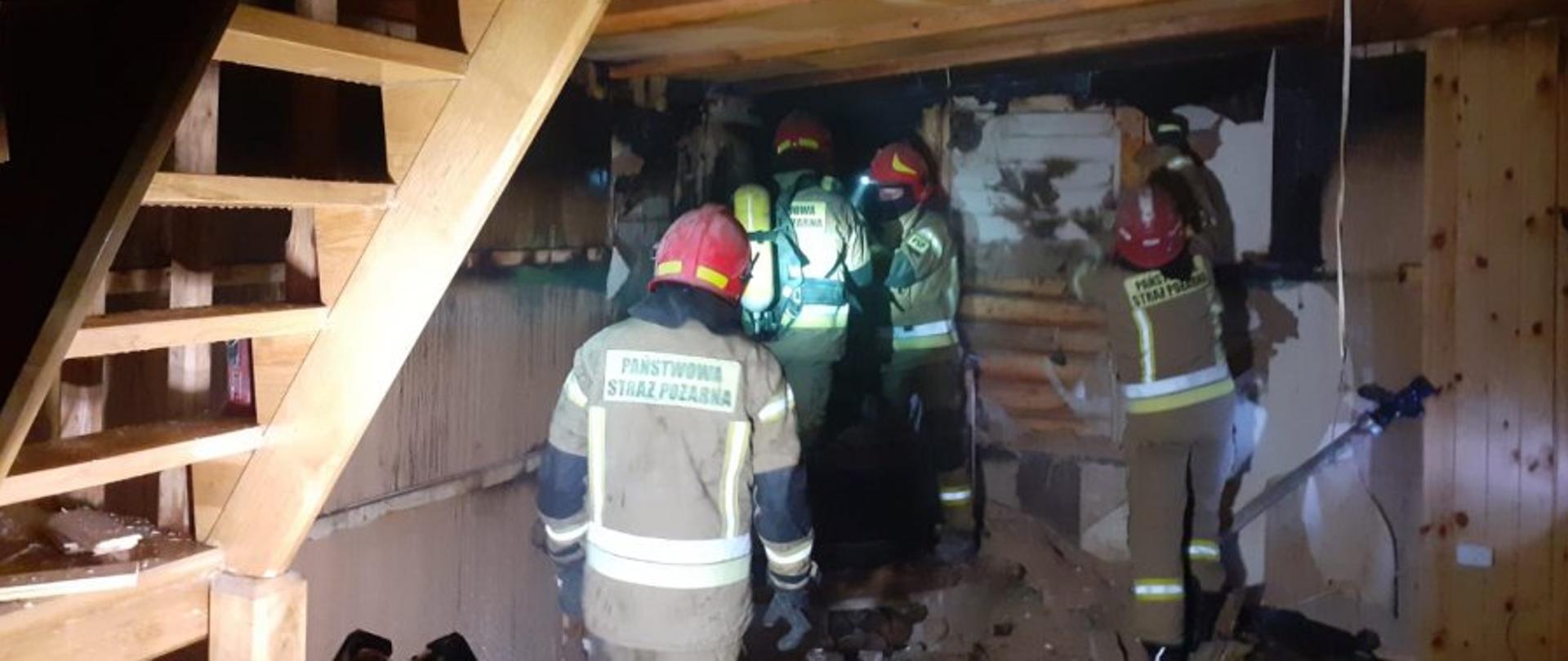 Zdjęcie zrobione w pomieszczeniu objętym pożarem. Na zdjęciu strażacy ubrani w ubrania specjalne usuwający skutki pożaru.