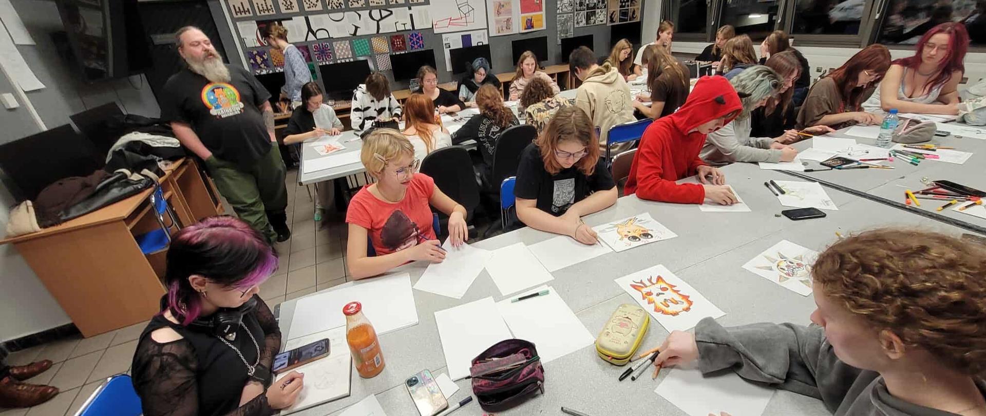 Dwa długie stoły z materiałami plastycznymi (kolorowe pisaki, kredki) oraz luźno rozłożonymi kartkami. Nad nimi pochylonych wielu młodych ludzi. W tle duże okna, za oknami ciemno. Na ścianach geometryczne kompozycje plastyczne. Z lewej strony stoi mężczyzna z długą siwą brodą