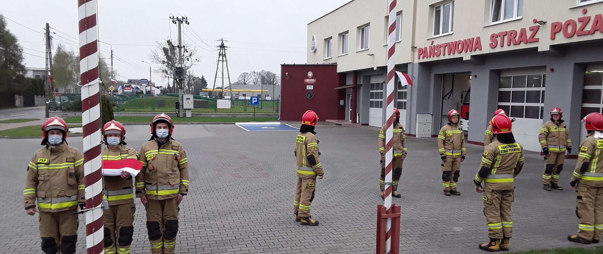 Zdjęcie przedstawia strażaków podczas wciągania flagi Polski na maszt, na dalszym planie widać innych strażaków podczas uroczystej zmiany służby