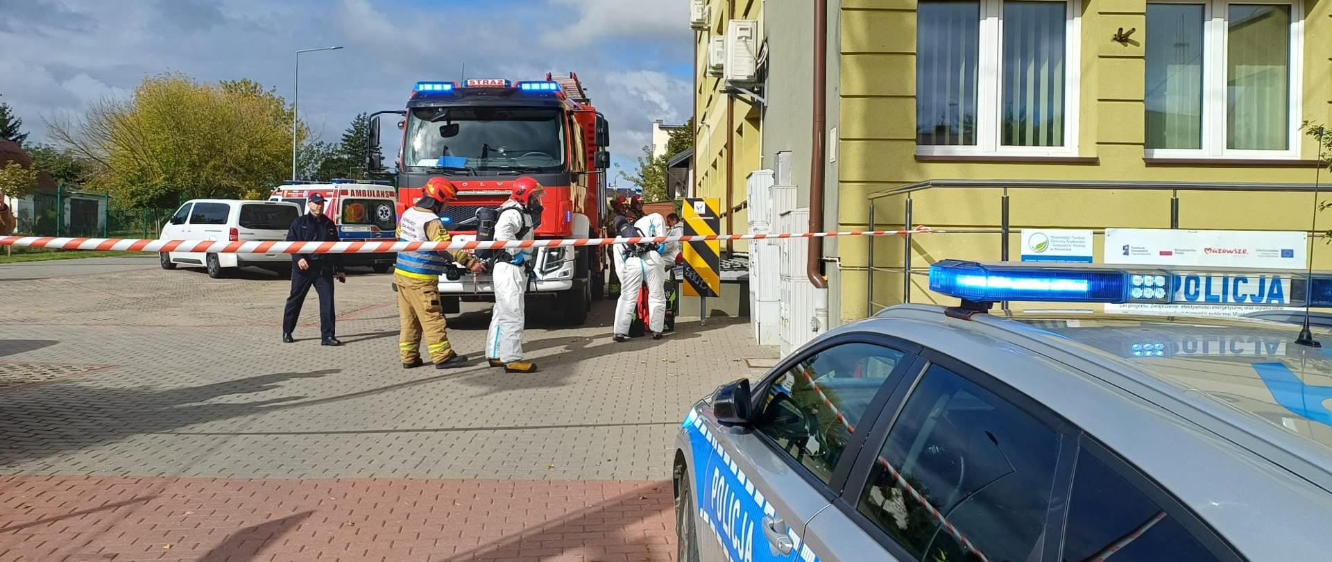 Na zdjęciu widać ćwiczenia służb PSP, Policji, Sanepidu oraz Urzędu Miasta. Służby stoją na placu, widać wóz strażacki, samochód policyjny. Plac odgrodzony jest białoczerwoną taśmą.