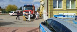 Na zdjęciu widać ćwiczenia służb PSP, Policji, Sanepidu oraz Urzędu Miasta. Służby stoją na placu, widać wóz strażacki, samochód policyjny. Plac odgrodzony jest białoczerwoną taśmą.