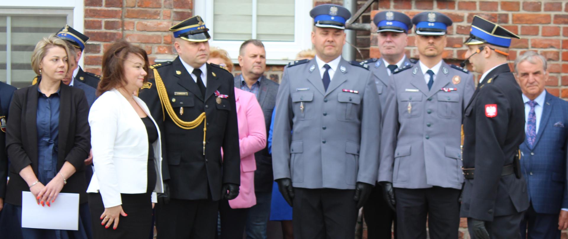 kobieta w jasnej marynarce i czarnej sukni stoi na baczność prtzed strażakiem. Obok kobiety stoi strażak, w tle grupa ludzi i policjanci
