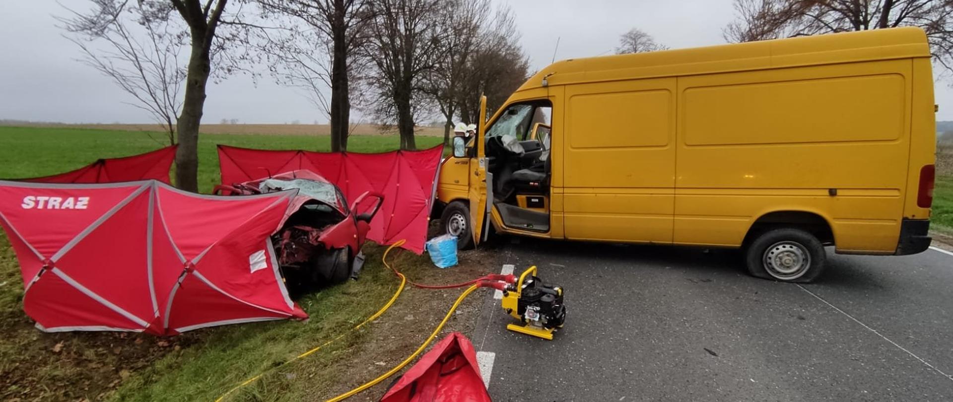 zdjęcie przedstawia rozstawione czerwone parawany z napisem straż dookoła rozbitego samochodu osobowego obok żółtego busa