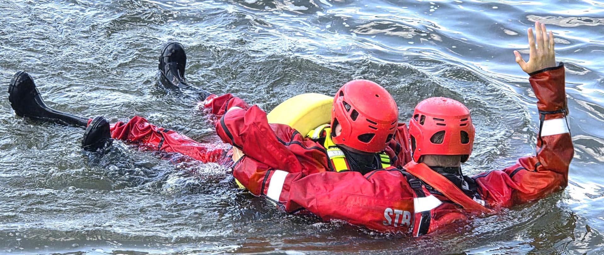 Zdjęcie przedstawia zbliżenie na kluczowy moment ćwiczeń – podjęcie i holowanie poszkodowanego w wodzie.
Oto szczegóły widoczne na fotografii:
Ratownik i poszkodowany: Widzimy dwóch ratowników w pełnym oporządzeniu (czerwone skafandry dry suit i czerwone kaski). Jedna osoba leży na plecach na wodzie (odgrywając rolę osoby ratowanej), a druga podtrzymuje ją i holuje.
Gesty i komunikacja: Ratownik udzielający pomocy ma podniesioną wysoko jedną rękę – jest to standardowy sygnał do załogi na łodzi/brzegu (np. sygnalizujący opanowanie sytuacji, gotowość do wyciągania lub prośbę o ściągnięcie liny).
Sprzęt: Pomiędzy ratownikami widoczny jest żółty pas ratowniczy (pas-węgorz / rescue tube), zapewniający dodatkową wyporność. Na skafandrze ratownika z prawej strony widoczny jest napis STRAŻ (od Straży Pożarnej).