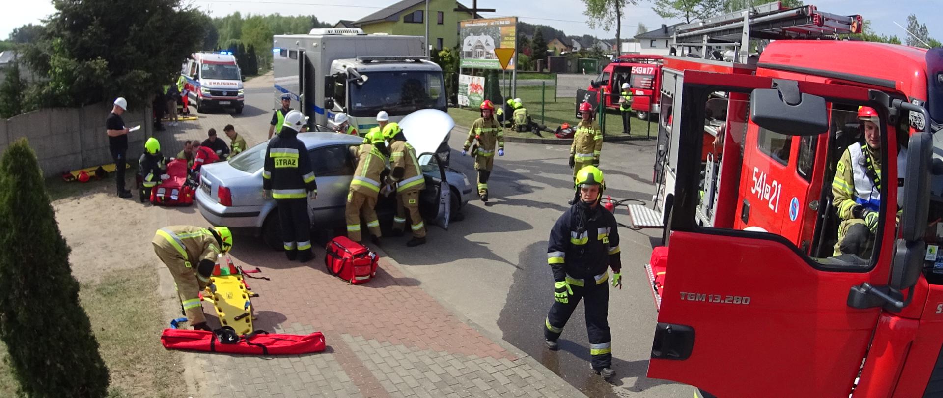 Strażacy w ubraniach specjalnych w trakcie uwalniania osoby poszkodowanej z rozbitego pojazdu. Za samochodem ratownicy udzielają pomocy. Obok stoi sędzia. Po prawej stronie samochody gaśnicze. W tle karetka pogotowia ratunkowego.
