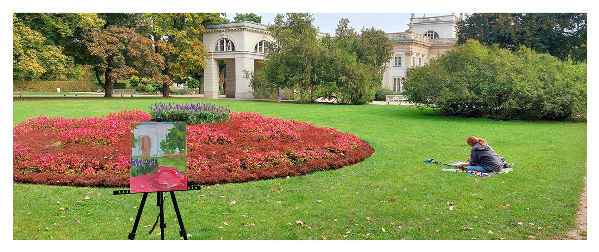 sztaluga z obrazem, w tle trawnik i pałac oraz siedząca na trawie malująca uczennica