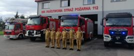 Strażacy oddający hołd w mundurach, na tle strażnicy oraz samochodów pożarniczych