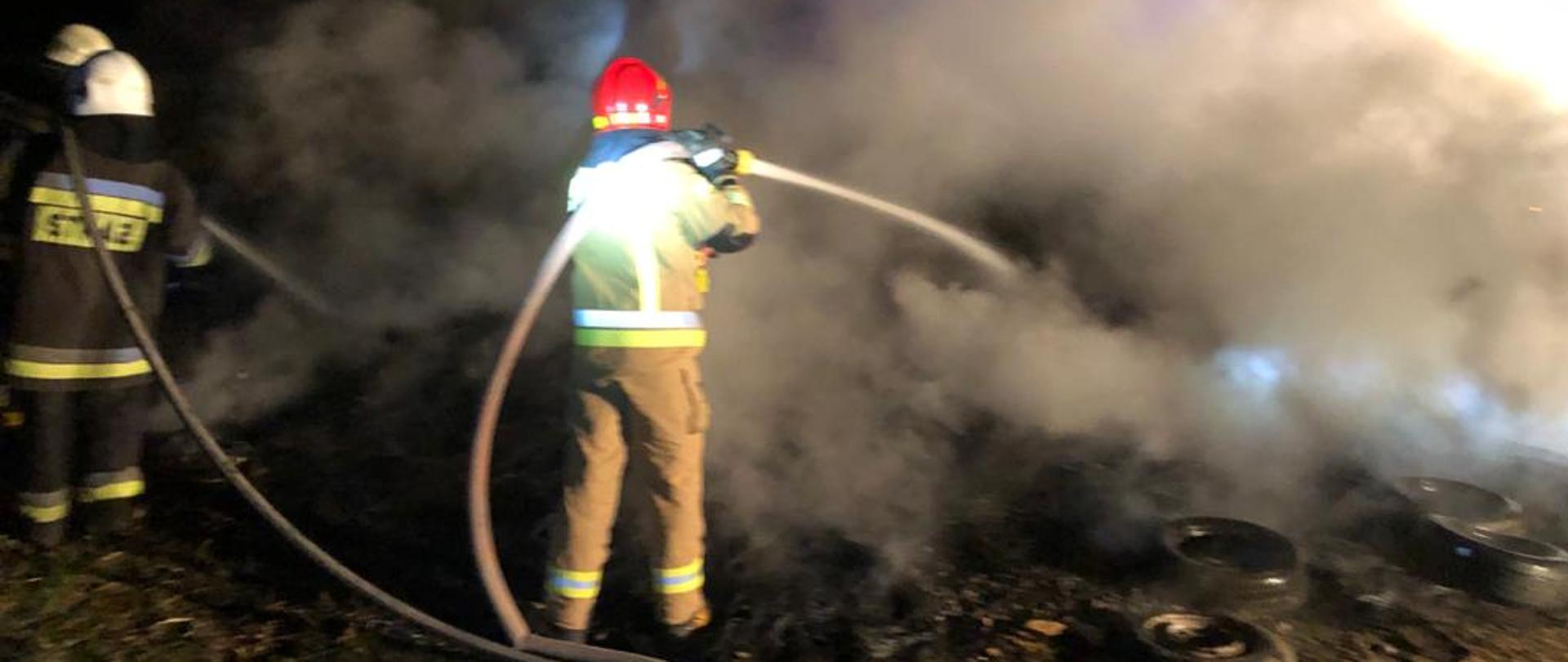 Na zdjęciu widoczni są strażacy w umundurowaniu specjalnym podający dwa prądy wody na palące się opony samochodowe. Widoczna jest duża ilość dymu wydostająca się z tlących opon.