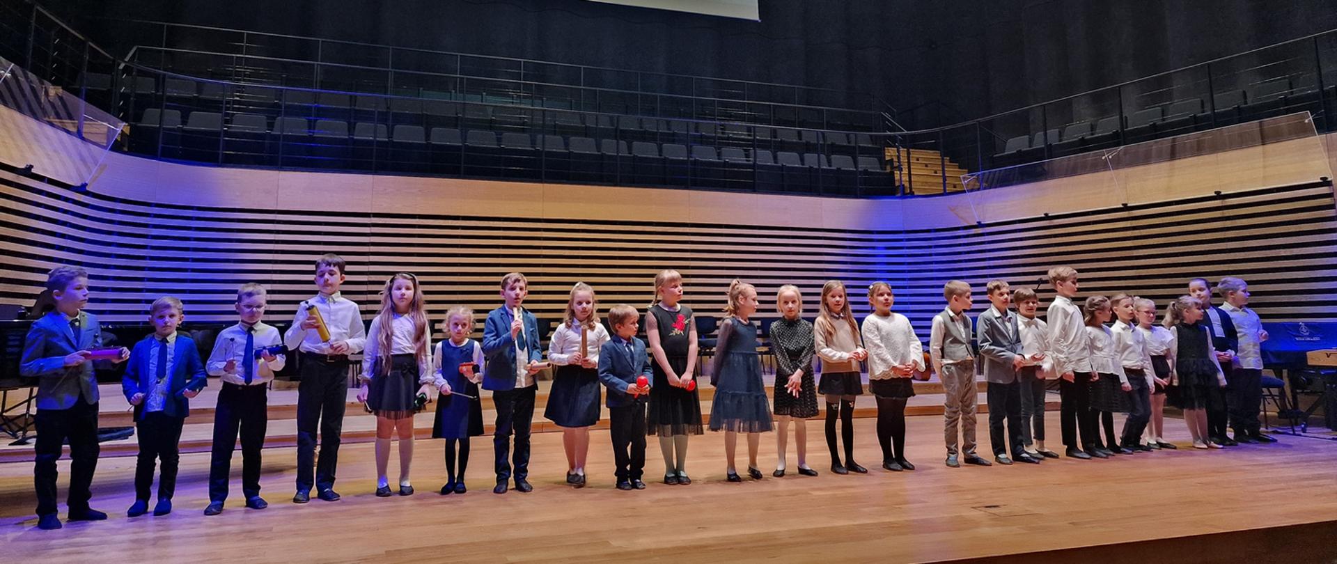 Dzieci stoją w rzędzie (24 osoby) na scenie sali koncertowej PSM w Jastrzębiu-Zdroju i śpiewają. Nad sceną widać rzutnik z napisem: klasa I c6 "Wiosna w błękitnej sukience" - przygotowanie mgr Aleksandra Musioł.