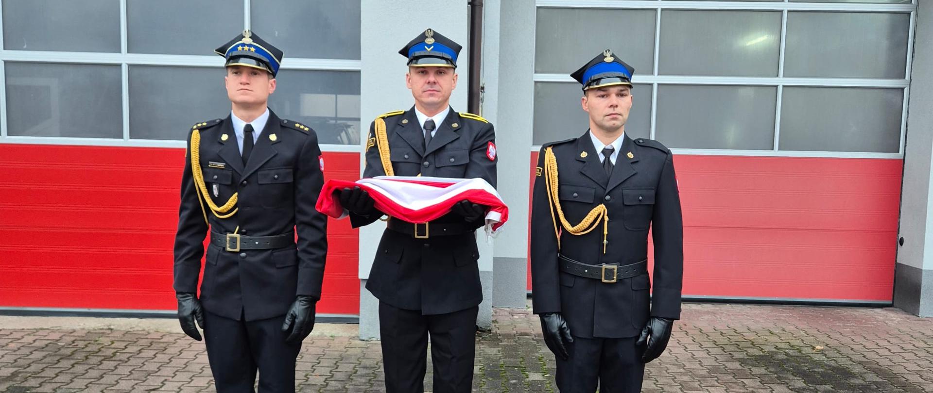 Trzech strażaków w galowym umundurowaniu PSP prezentuje złożoną biało-czerwoną flagę państwową podczas uroczystości 11 listopada.
