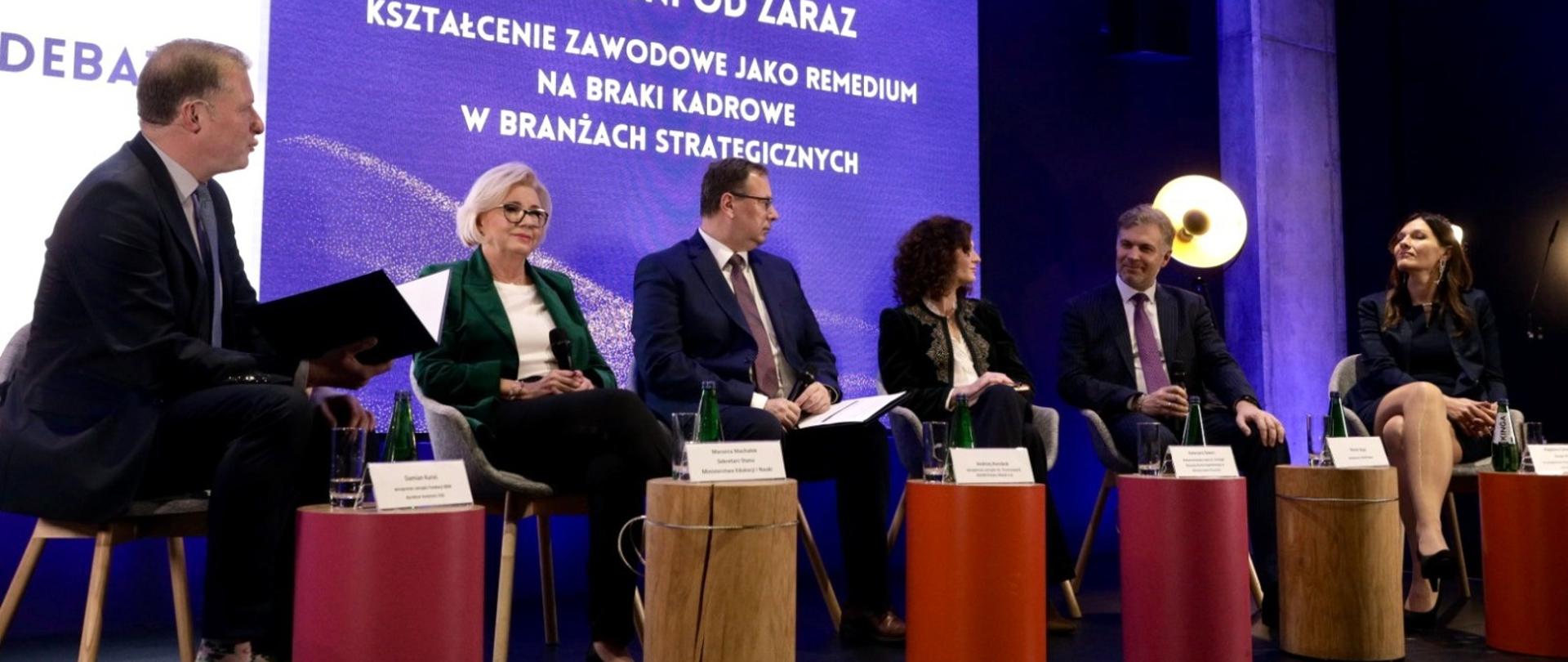 Na tle fioletowej ściany z napisem Zawodowcy potrzebni od zaraz siedzi grupa sześciu osób, przed nimi małe okrągłe stoliki.
