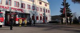 Strażacy przed jednostką na tle garażu z samochodami pożarniczymi oraz powiewająca flaga państwowa na maszcie 