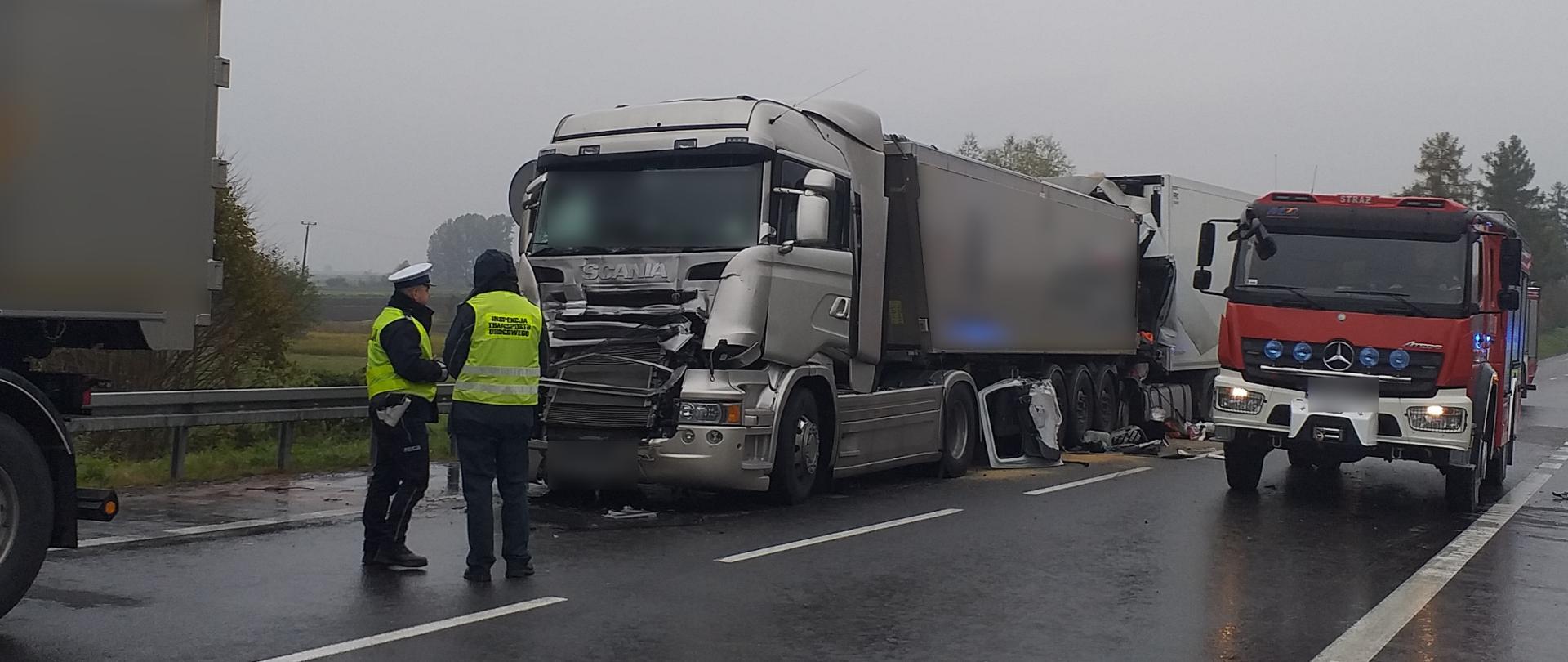 Droga jedno jezdniowa o dwóch pasach ruchu. Przed rozbitą ciężarówką ze srebrną kabiną stoi inspektor ITD i policjant. Za ciężarówką stoi inna ciężarówka ze zmiażdżoną kabiną. Obok wóz strażacki. 