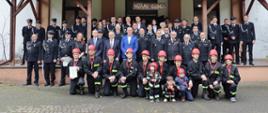 zdjęcie pamiątkowe kilkudziesięciu uczestników spotkania w miejscowości Dzierzgów na pierwszym planie młodzieżówka osp w czerwonych kaskach na głowie ubrana w umundurowanie koszarowe osp na drugim planie zaproszeni oficjele na planie trzecim stoją goście i druhowie ochotniczych straży pożarnych w mundurach galowych, w tle budynek z napisem Witamy Gości