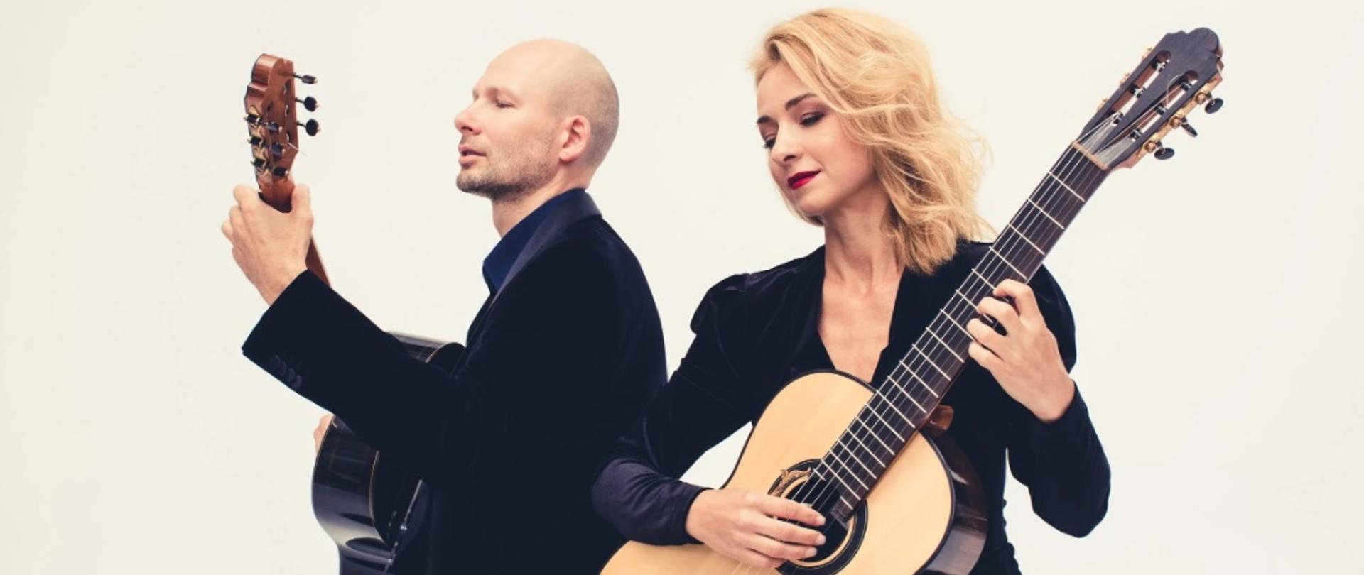 Na zdjęciu widać dwie osoby siedzące tyłem do siebie, grające na gitarach klasycznych. Kobieta po prawej stronie ma blond włosy, jest elegancko ubrana w ciemny strój i skupiona na grze. Mężczyzna po lewej stronie ma krótkie włosy, również ubrany jest w ciemny garnitur i gra na gitarze. Tło jest jasne i jednolite, co podkreśla postacie i instrumenty, nadając zdjęciu elegancki, artystyczny charakter.