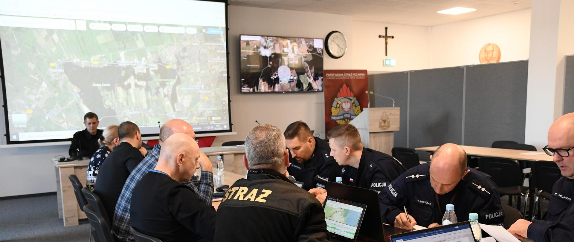 Funkcjonariusze służb mundurowych oraz instytucji siedzą za stołem przed swoimi laptopami niedaleko siedzi za stołem strażak za nim jest monitor na którym wyświetlana jest obecna sytuacja oraz telewizor z prognozami rozwoju sytuacji.