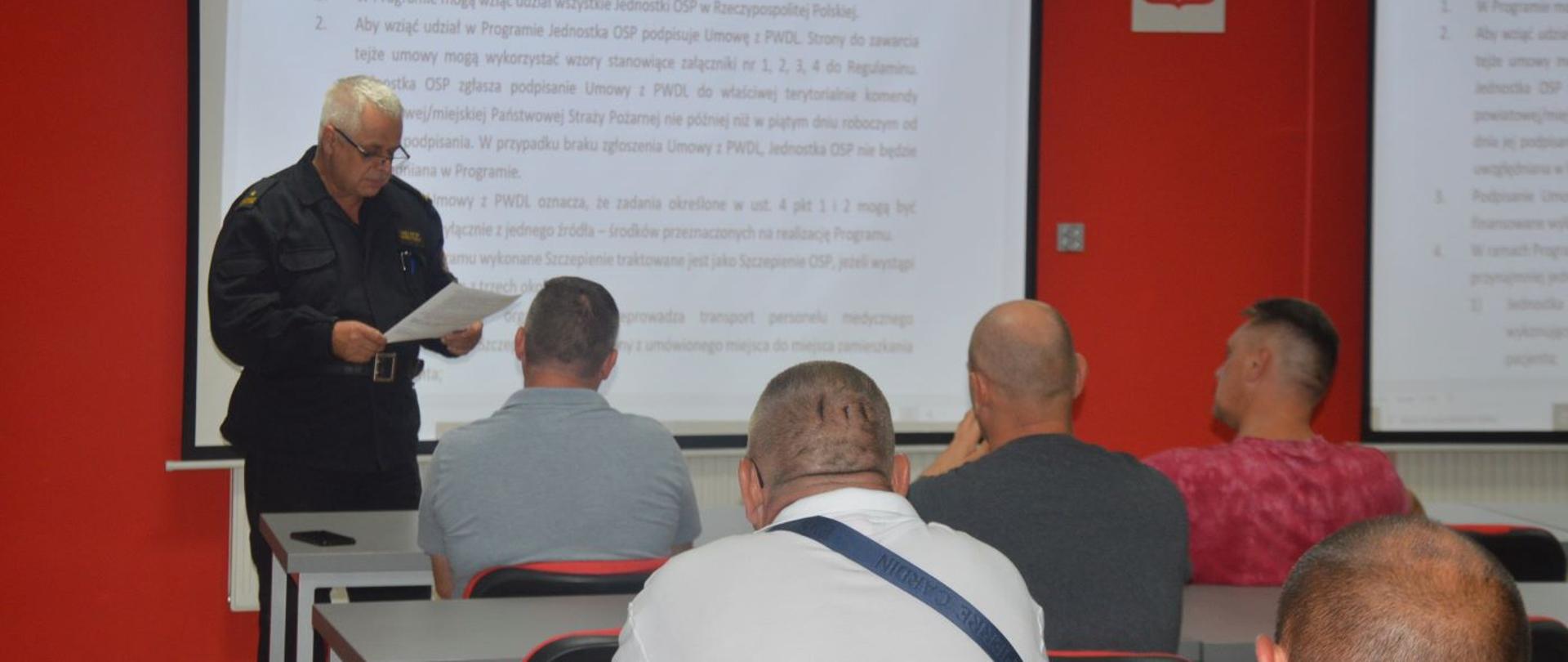 Świetlica Komendy Powiatowej PSP w Rawiczu. Zastępca komendanta powiatowego PSP, który prowadzi spotkanie ubrany jest w mundur dowódczo-sztabowy. W rękach trzyma dokument, z którym zapoznaje uczestników narady. Uczestniczący w naradzie siedzą na krzesłach, przy stołach, zwróceni w stronę ekranów, na których wyświetlane są zasady programu #SzczepimySię z OSP.
