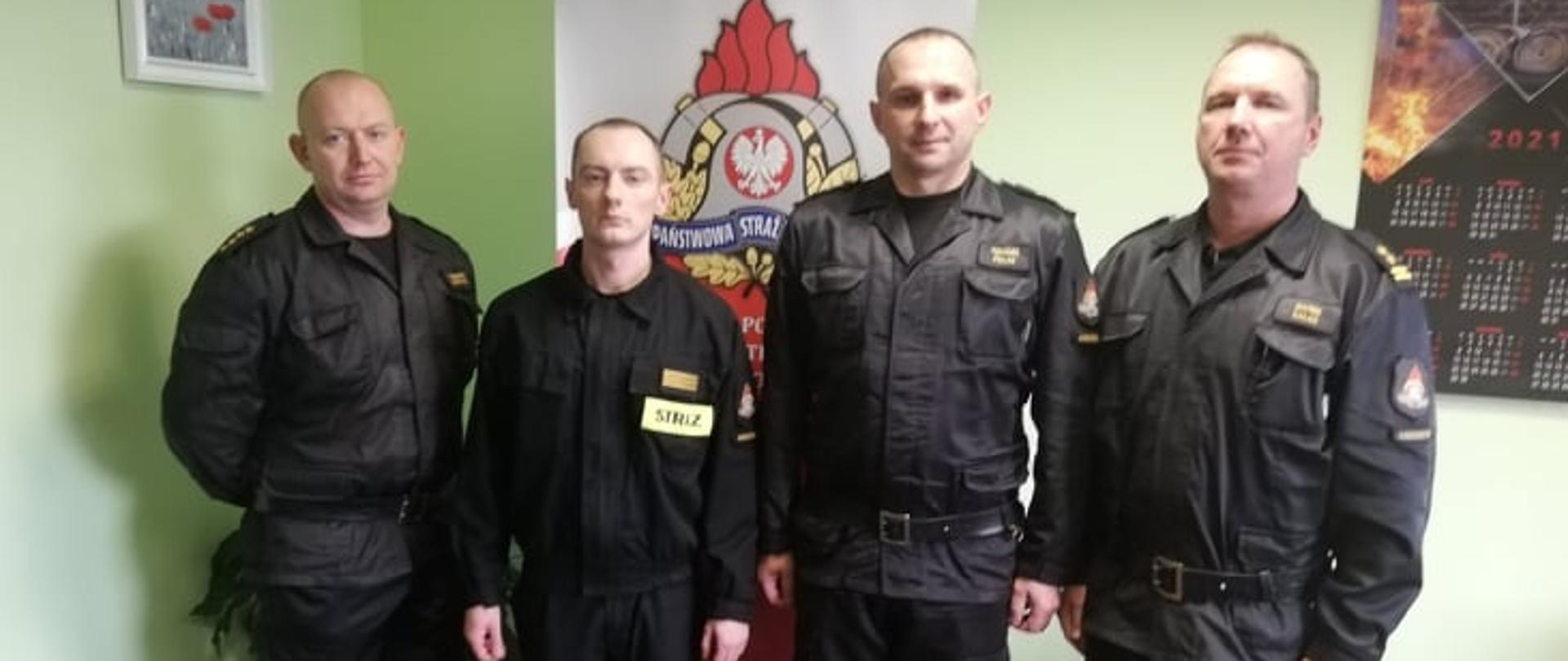 W gabinecie komendanta powiatowego PSP komendant powiatowy wraz ze swoim zastępcą w obecności z-cy dowódcy jednostki ratowniczo - gaśniczej złożył gratulację strażakowi, który w czasie wolnym od służby udaremnił próbę samobójczą. Wymienione osoby stoją w szeregu na tle baneru komendy powiatowej