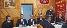 Zastępca Komendanta Powiatowego PSP Skarżysko-Kamienna przemawia podczas zebrania sprawozdawczego w OSP Łączna. Po jego lewej jak stronie siedzi prezes OSP, a po prawej wójt gminy, wiceminister spraw zagranicznych , radny powiatowy i naczelnik OSP.