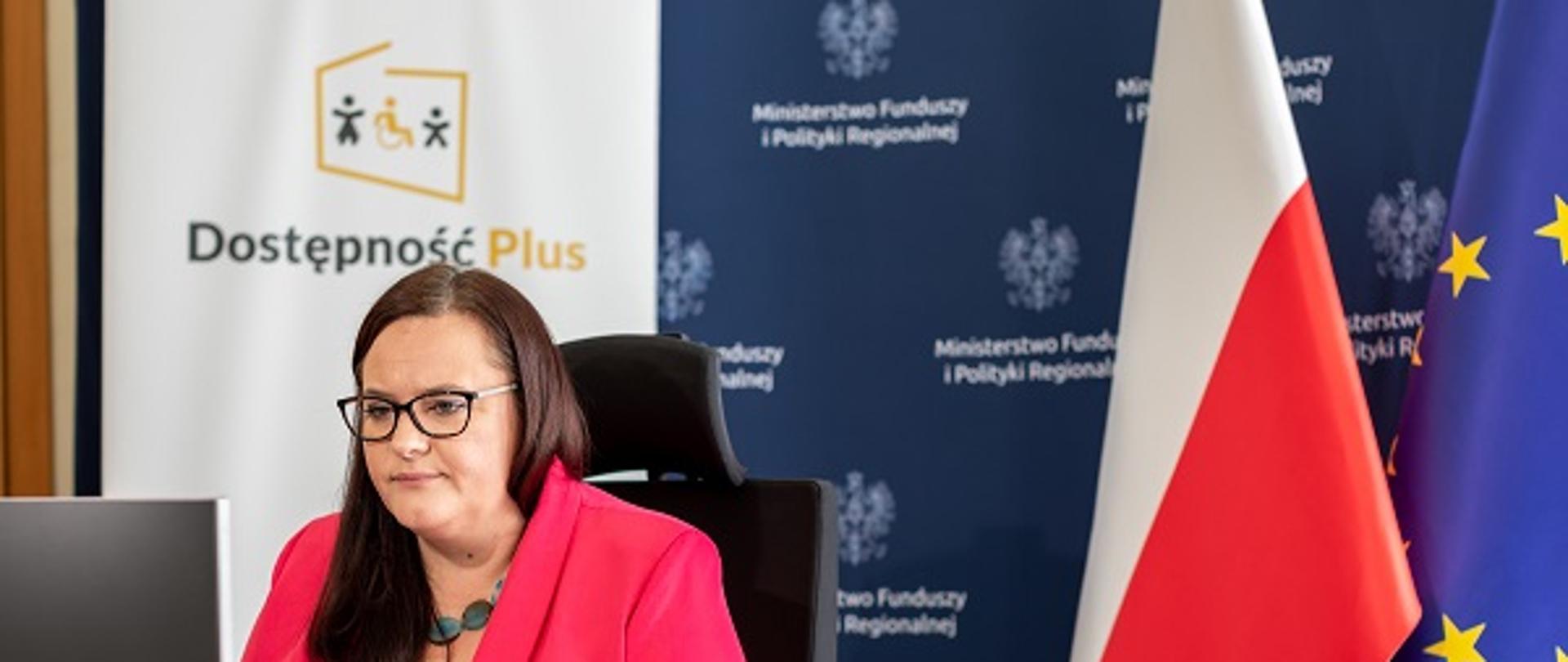 Minister Małgorzata Jarosińska-Jedynak siedzi przy biurku przed komputerem. Za plecami ścianka z logo Programu Dostępność Plus oraz MFiPR, a także flagi Polski i UE.
