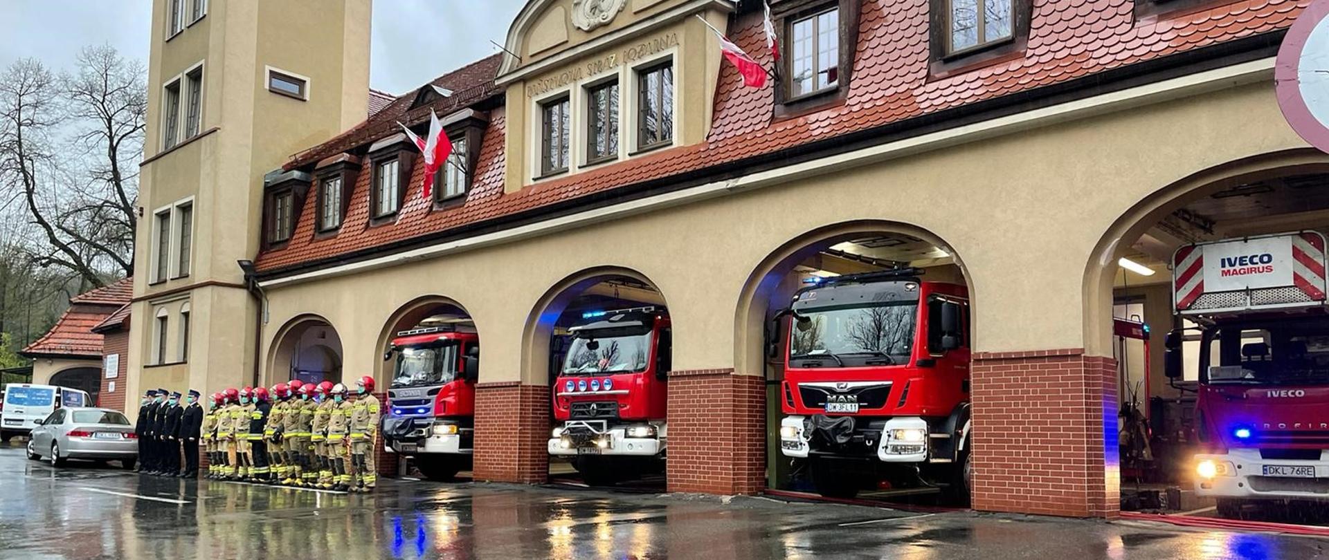 Widok na plac Komendy Powiatowej Straży Pożarnej w Kłodzku. Uroczysta zbiórka funkcjonariuszy. W tle otwarte bramy garażowe z samochodami ratowniczo-gaśniczymi.