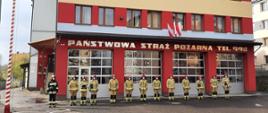 Kolorowe zdjęcie przedstawia uroczystą zbiórkę strażaków w ubraniach bojowych na placu przed budynkiem Państwowej Straży Pożarnej w Brzozowie.
